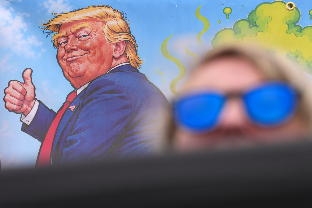 A protester wears sunglasses and sits in front of a poster depicting U.S. President Donald Trump during his visit to the Irish Open at the Trump International Golf Links course in Doonbeg, Ireland, Sunday, Sept. 13, 2026.(AP Photo/Kin Cheung)