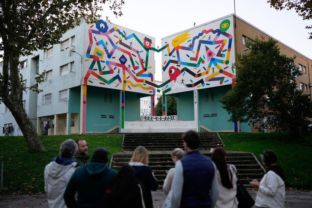 Foreign university students on a tour take notes about a mural by artist Bernardo P Carvalho, inspired by the United Nations Sustainable Development Goal 11, Sustainable Cities and Communities, at the housing project Zambujal in Amadora, on the outskirts of the Portuguese capital Lisbon, Tuesday, Nov. 18, 2025. (AP Photo/Armando Franca)