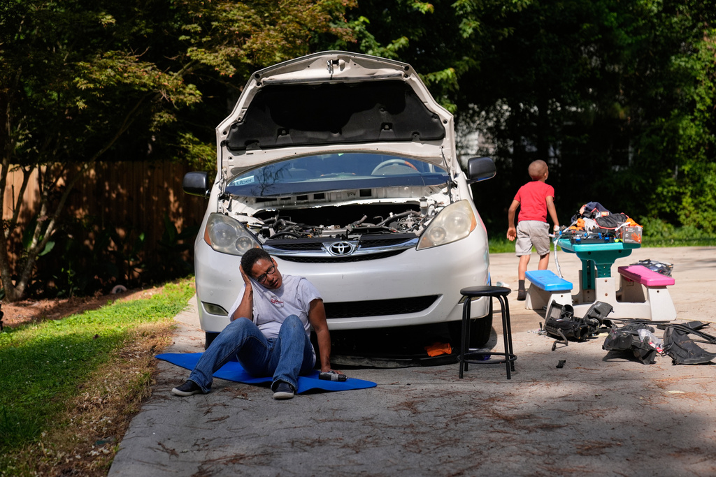 Sechita McNair, center, rests in the summer heat as she repairs her family van, while her adopted son, Derrick McNair-White, right, plays in the driveway of their rental home on June 6, 2025, in Jonesboro, Ga. (AP Photo/Brynn Anderson)