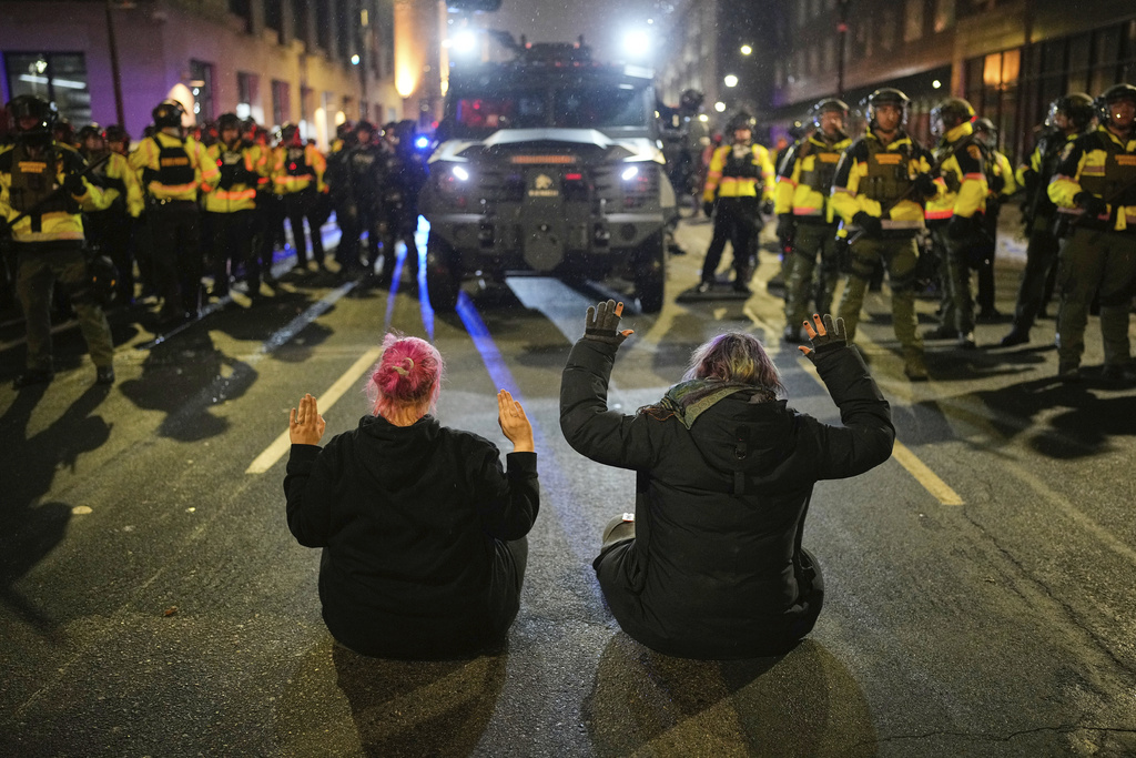 Two people sit in the street with their hands up in front of Minnesota State Patrol during a protest and noise demonstration calling for an end to federal immigration enforcement operations in the city, Friday, Jan. 9, 2026, in Minneapolis. (AP Photo/John Locher)