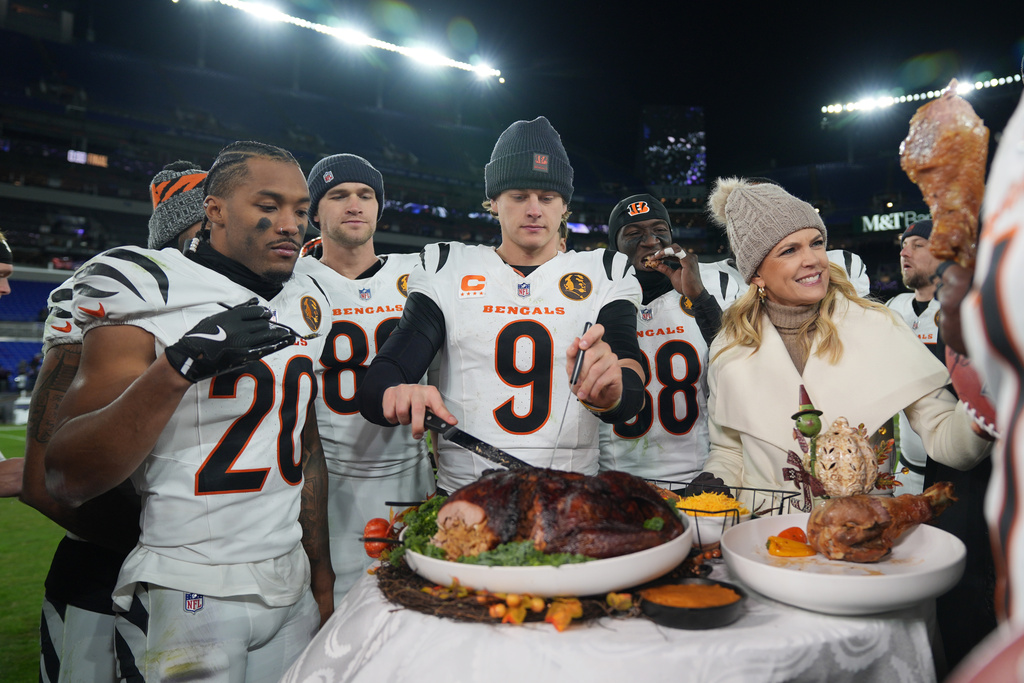 Cincinnati Bengals quarterback Joe Burrow (9) joined by NBC Sports sideline reporter Melissa Stark, right, teammates DJ Turner II (20), Mike Gesicki (88), and DJ Ivey (38) carves a turkey after NFL football game against the Baltimore Ravens, Thursday, Nov. 27, 2025, in Baltimore. (AP Photo/Stephanie Scarbrough)