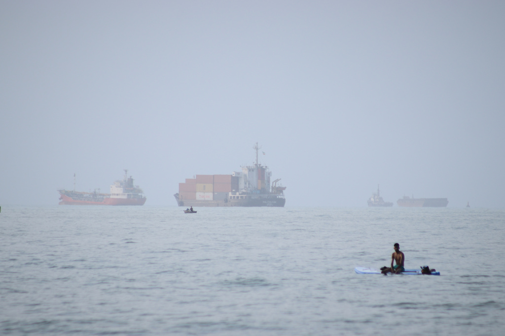 FILE - A man sits on a surfboard as a container ship and other commercial vessels appear anchored in the Strait of Hormuz off Bandar Abbas, Iran, Monday, July 27, 2026. (Razieh Poudat/ISNA via AP, File)