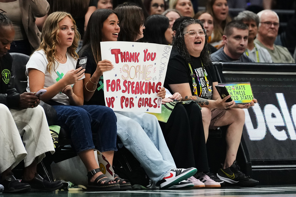 A fan holds a sign for Indiana Fever guard Sophie Cunningham in reference to her comments on transgender athletes as a fan wearing Seattle Storm gear holds a competing sign during the first half of a WNBA basketball game, Tuesday, July 28, 2026, in Seattle. (AP Photo/Lindsey Wasson)
