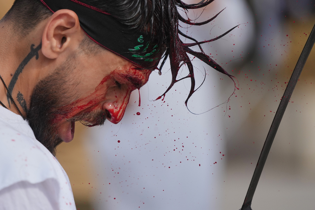 EDS NOTE: GRAPHIC CONTENT - A man bleeds from a ceremonial head wound as he strikes his head during Ashoura, when Shiites commemorate the martyrdom of Imam Hussein, the grandson of the Prophet Muhammad, amid buildings destroyed by Israeli strikes in Nabatiyeh, southern Lebanon, Friday, June 26, 2026. (AP Photo/Hassan Ammar)