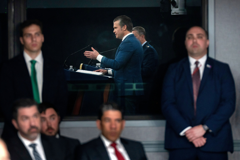 Staff members watch as Defense Secretary Pete Hegseth, reflected in a window, speaks during a press briefing at the Pentagon, Monday, March 2, 2026, in Washington. (AP Photo/Mark Schiefelbein)