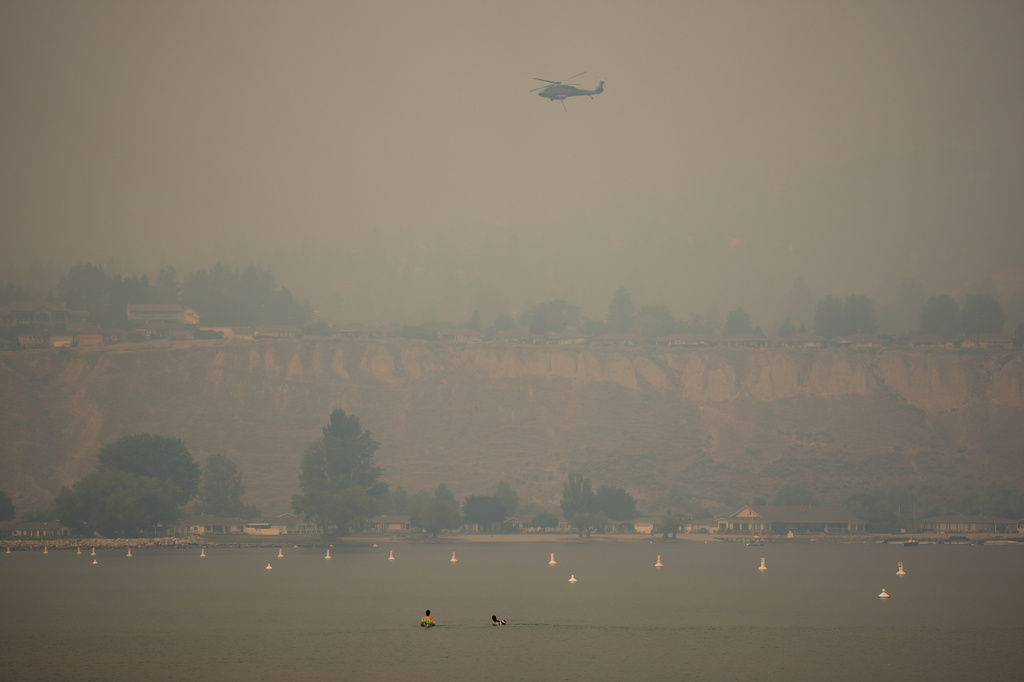 People float in the water while a helicopter being used to battle wildfires flies overhead as smoke from the Bald Range wildfire envelops Okanagan Lake, in Penticton, B.C., on Sunday, Aug. 9, 2026. (Darryl Dyck/The Canadian Press via AP)