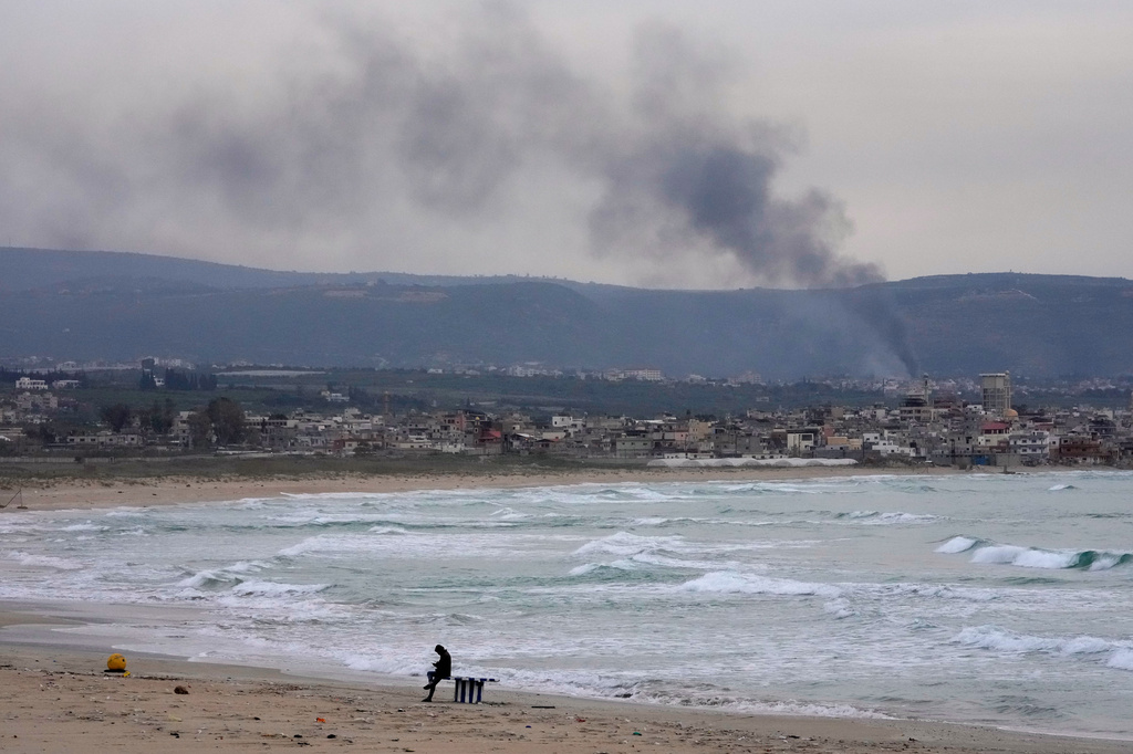 A man checks his mobile phone on a public beach, as smoke, background, rises from Israeli artillery shells on Qlaileh village, as it s seen from Tyre city, south Lebanon, Saturday, March 28, 2026. (AP Photo/Hussein Malla)