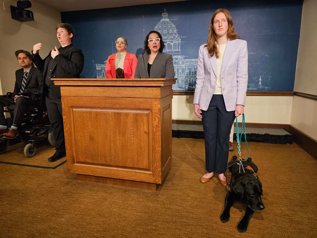 Minnesota Human Rights Commissioner Rebecca Lucero, at the podium, holds a news conference at the State Capitol in St. Paul on Wednesday, March 11, 2026, to announce a settlement with the ride-sharing company Lyft that will ensure the rights of blind and other disabled passengers across the country to travel with their service animals. College student Tori Andres, right, turned to the Minnesota Department of Human Rights after several Lyft drivers refused to let her service dog, Alfred, ride along with her. (AP Photo/Steve Karnowski)