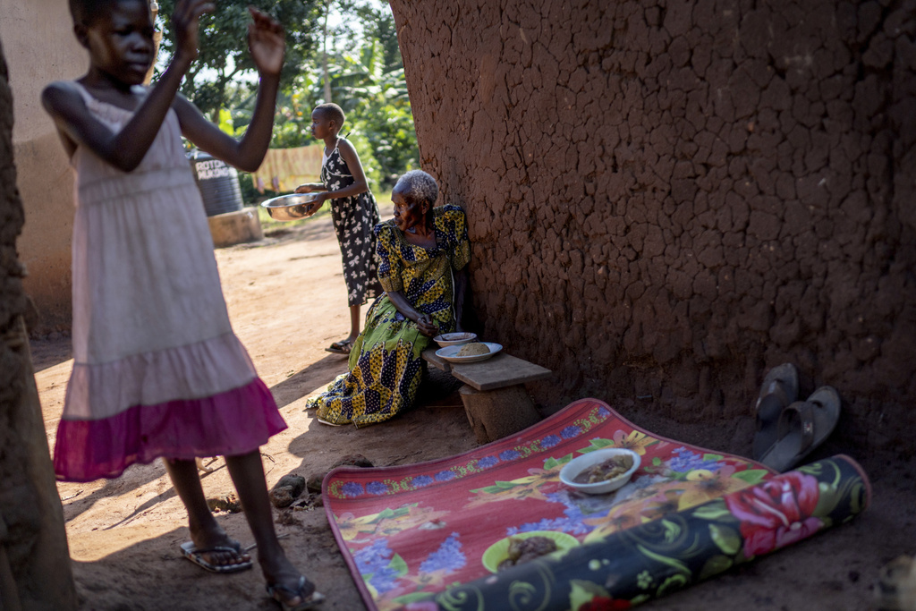 Rose Liru, 94, right, eats dinner prepared by her granddaughter, Parvin Nakawesi, 9, left, and grandniece, Brenda Mungulu, 11, rear, at the home they share, Friday, Nov. 22, 2024, in Magogo, Uganda. (AP Photo/David Goldman)