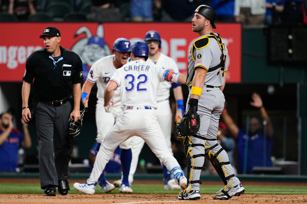 Pittsburgh Pirates catcher Joey Bart, right, and umpire Dan Merzel, left, look to the outfield as Texas Rangers' Evan Carter (32) and others celebrate Carter's inside the park home run in the third inning of a baseball game Thursday, April 23, 2026, in Arlington, Texas. (AP Photo/Tony Gutierrez)