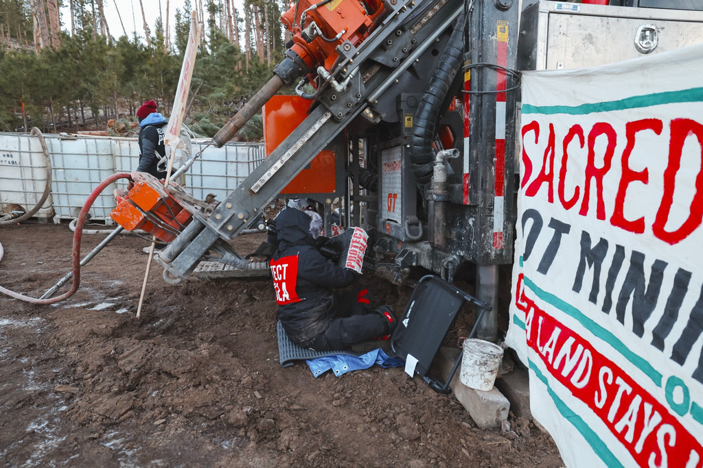 A demonstrator in a jacket with the words Protect Pe'Sla sits on the ground near drilling equipment on Thursday, April 30, 2026, in Pennington County, S.D., with a banner reading Sacred ground not mining bound. (Angel White Eyes via AP)
