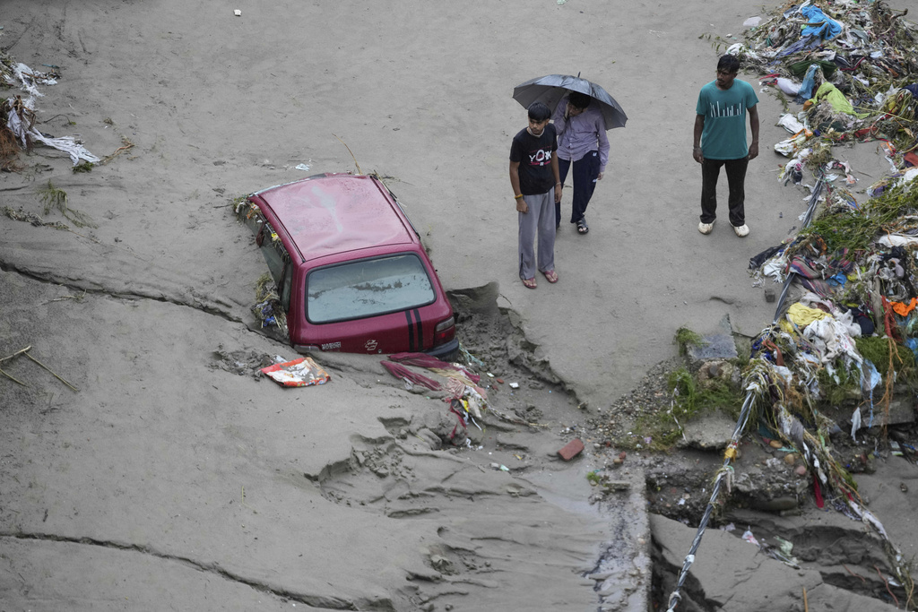 People stand next to a stuck car in the aftermath of flash floods at the banks of Tawi river in Jammu, India, Wednesday, Aug. 27, 2025. (AP Photo/Channi Anand)