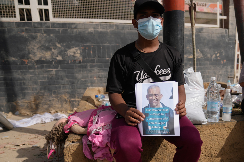 Kevin Tovar sits near his damaged home holding leaflets with a photo of his missing father in La Guaira, Venezuela, Friday, June 26, 2026, two days after twin earthquakes struck the country. (AP Photo/Ariana Cubillos)