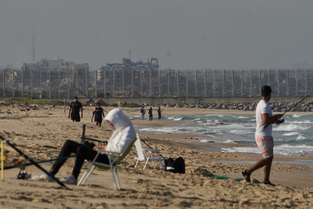 Backdropped by buildings destroyed during Israeli ground and air operations in the northern Gaza Strip, people walk and fish at Zikim Beach, near Ashkelon, in southern Israel, Friday, Oct. 17, 2025. (AP Photo/Leo Correa)