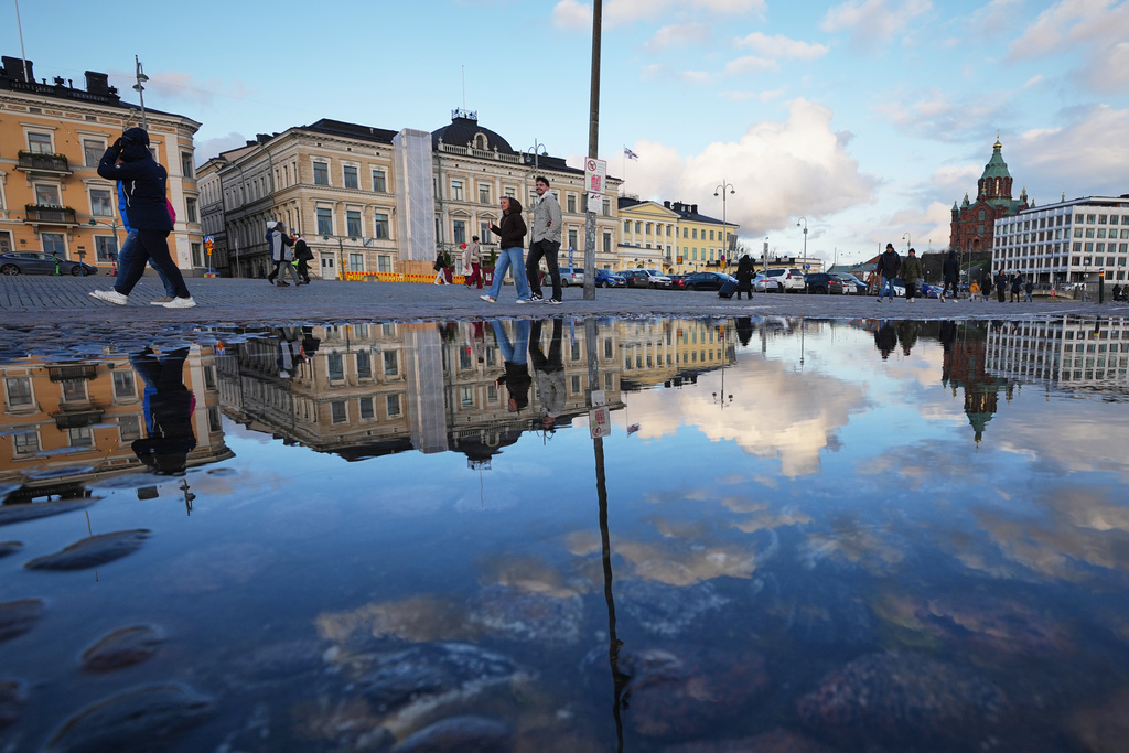 People walking along the square are reflected in a puddle in Helsinki, Finland, Friday, Nov. 14, 2025. (AP Photo/Sergei Grits)
