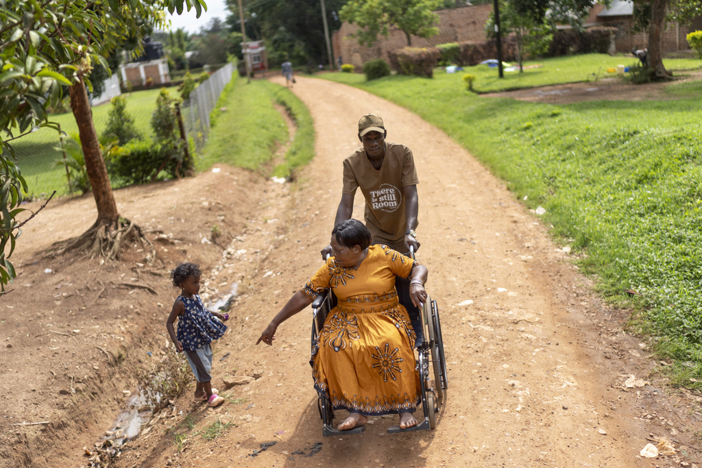 Norah Kasozi, 73, gestures to her granddaughter, Shalom, 4, to be careful near a ditch as she is pushed in a wheelchair to her home by Isaac Okware following a physical therapy visit to a nearby clinic, Saturday, Nov. 16, 2024, in Mukono, Uganda. (AP Photo/David Goldman)