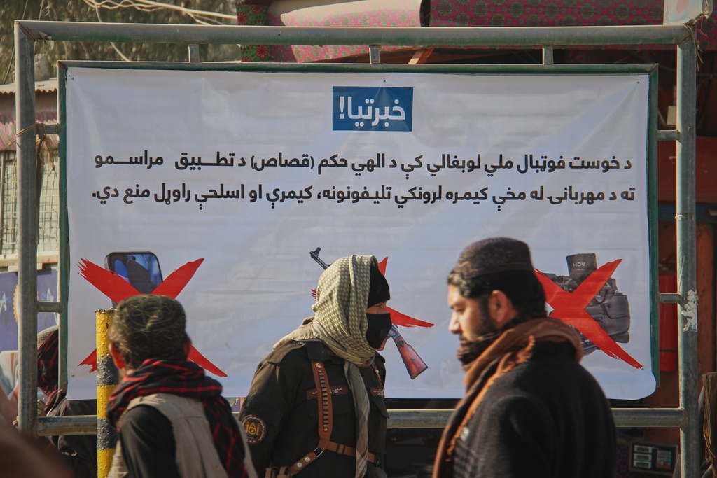 A Taliban policeman, center, and two other men walk past a poster stating that cameras, phones, and weapons are banned inside a stadium where a public execution was taking place in the city of Khost, eastern Afghanistan, Tuesday, Dec. 2, 2025. (AP Photo/Saifullah Zahir)