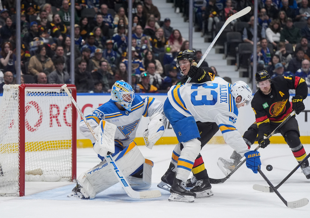 St. Louis Blues' Logan Mailloux (23) checks Vancouver Canucks' Aatu Raty (54) in front of St. Louis goalie Jordan Binnington (50) while Vancouver's Brock Boeser (6) tries to get his stick on the puck during the third period of an NHL hockey game in Vancouver, Saturday, March 21, 2026. (Darryl Dyck/The Canadian Press via AP)