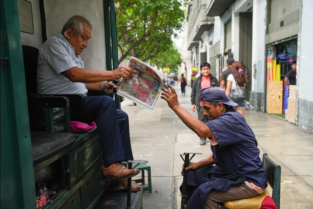 A shoe shiner passes a newspaper to a client near the government palace, the office of the president, in Lima, Peru, Wednesday, Feb. 18, 2026, the day after lawmakers voted to remove interim President Jose Jeri from office as he faces corruption allegations. (AP Photo/Guadalupe Pardo)