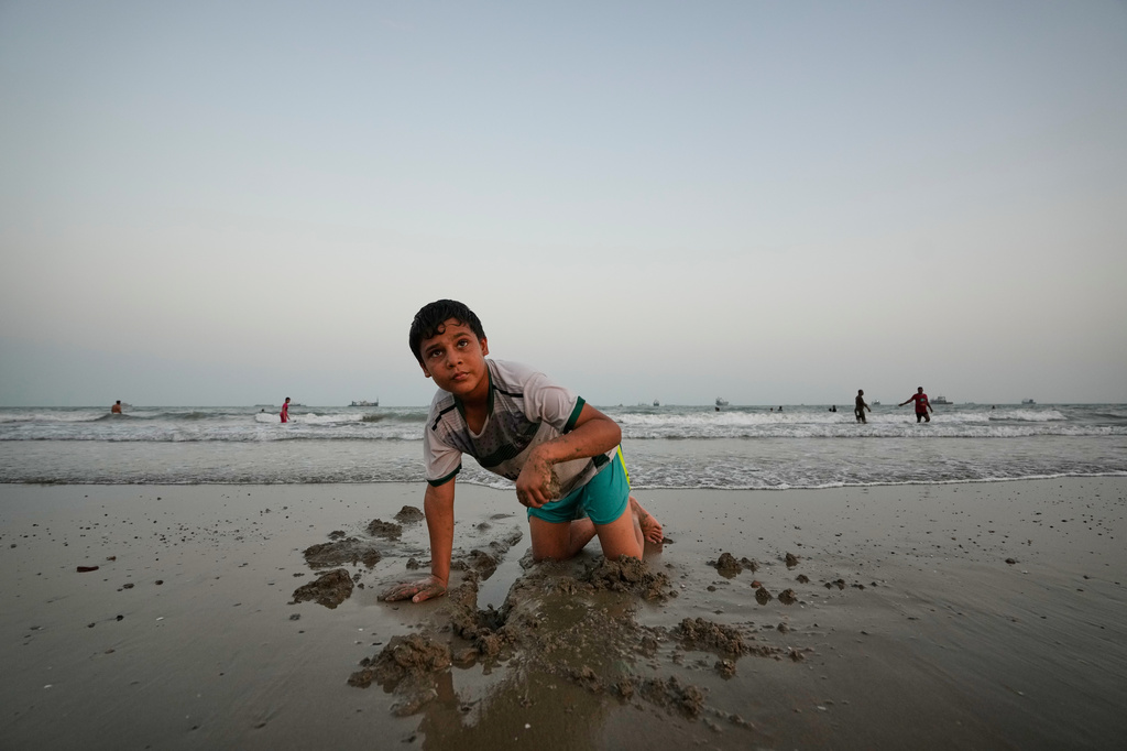 A boy plays in wet sand as others swim and commercial vessels lie anchored in the Strait of Hormuz off Bandar Abbas, Iran, Friday, Sept. 4, 2026. (AP Photo/Vahid Salemi)