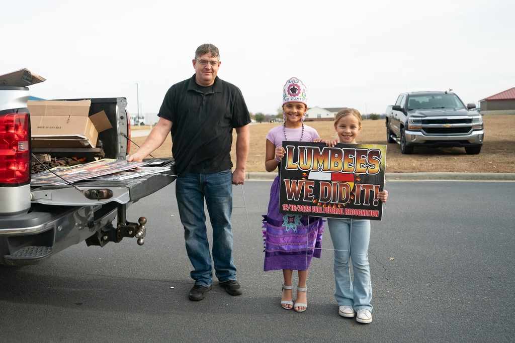 Children pose for a photo with a man selling signs after passage of the National Defense Authorization Act by the U.S. Senate, during a watch party hosted by the Lumbee Tribe of North Carolina, Wednesday, Dec. 17, 2025, in Pembroke, N.C. (AP Photo/Allison Joyce)