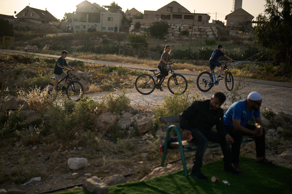 Young Israeli settlers ride bicycles past Palestinian men sitting near a small soccer pitch in the West Bank Bedouin village of Umm al-Khair, Tuesday, May 12, 2026. (AP Photo/Leo Correa)