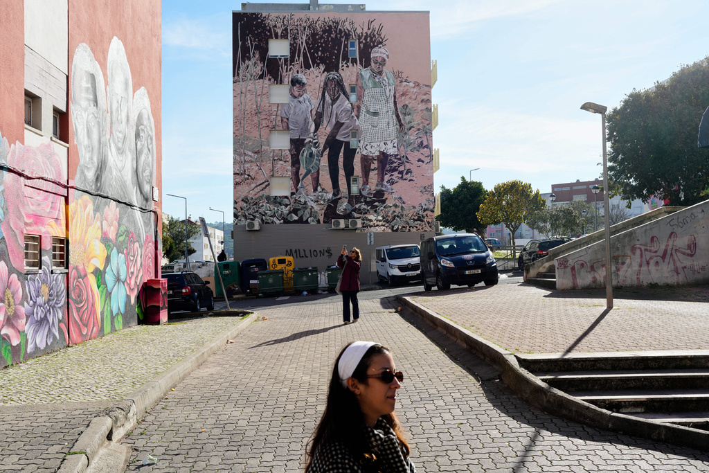 Tour guide Ines, in the foreground, listens to comments by visitors about the mural by artist Ligia Fernandes, inspired by the United Nations Sustainable Development Goal 2, Zero Hunger, in the background, at the housing project Zambujal in Amadora, on the outskirts of the Portuguese capital Lisbon, Thursday, Nov. 27, 2025. (AP Photo/Armando Franca)