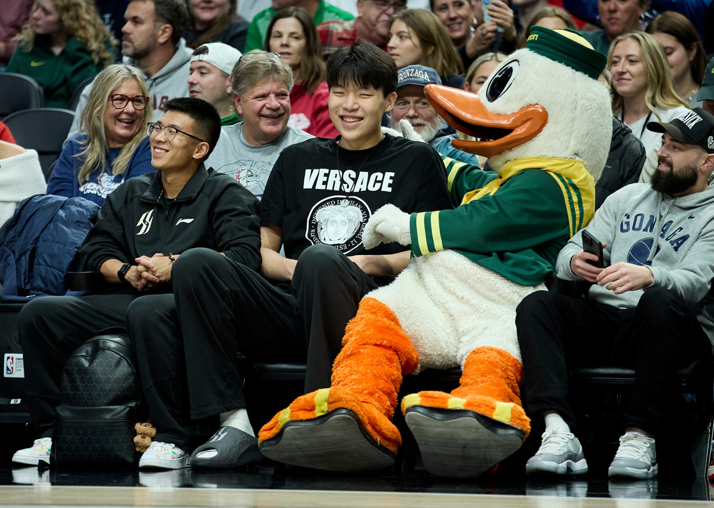 The Oregon mascot, second from right, sits next to Portland Trail Blazers center Yang Hansen, center, during the first half of an NCAA college basketball game between Oregon and Gonzaga in Portland, Ore., Sunday, Dec. 21, 2025. (AP Photo/Craig Mitchelldyer)