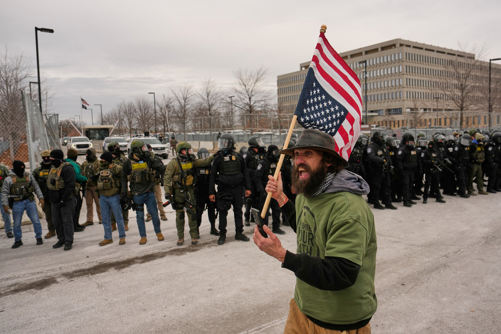 A protester carries an upside down American flag in front of federal immigration officers outside Bishop Henry Whipple Federal Building, Thursday, Jan. 15, 2026, in Minneapolis. (AP Photo/John Locher)
