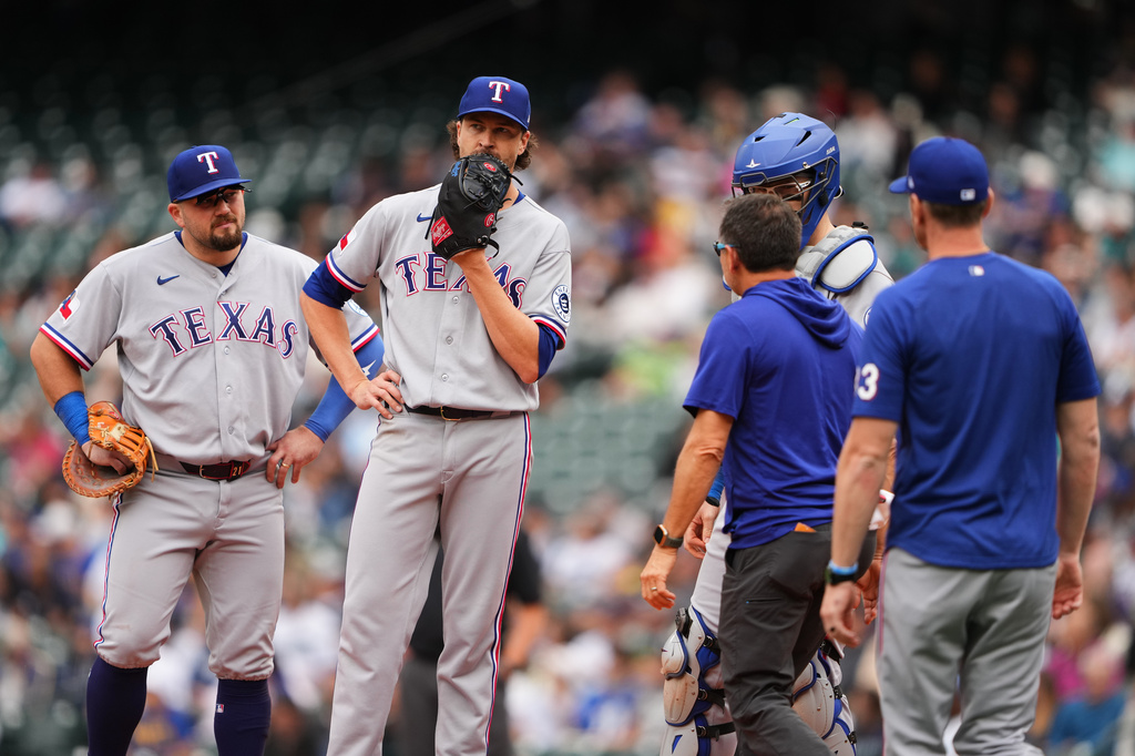 A trainer comes to check on Texas Rangers starting pitcher Jacob deGrom, center, after Seattle Mariners' Cole Young hit a line drive for a single straight at deGrom during the sixth inning of a baseball game, Thursday, Sept. 10, 2026, in Seattle. (AP Photo/Lindsey Wasson)