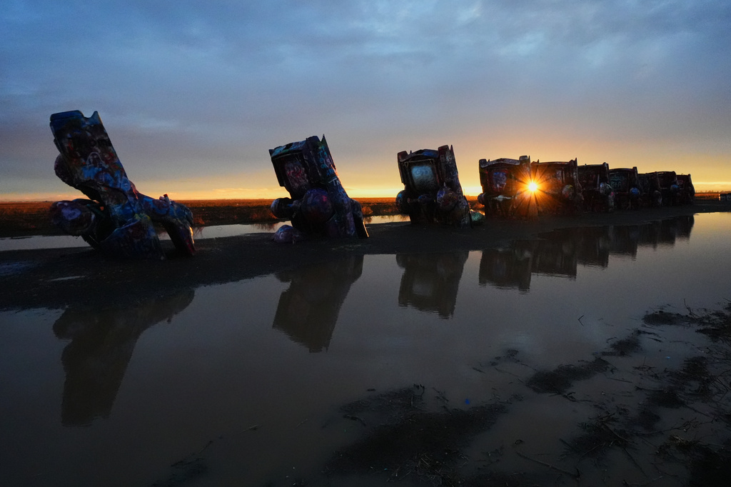 A puddle from a midday storm surrounds vehicles as the setting sun peeks through at Cadillac Ranch, a roadside attraction along Route 66 in Amarillo, Texas, Thursday, Nov. 20, 2025. (AP Photo/Julio Cortez)