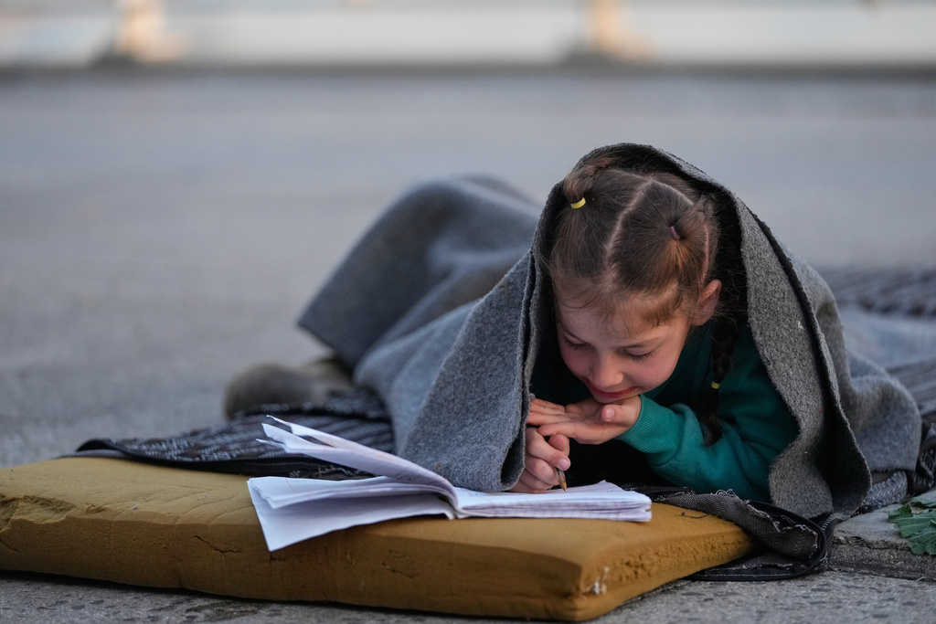 Hanaa, 7, displaced by Israeli airstrikes in Dahiyeh, Beirut's southern suburbs, writes in a notebook while wrapped in a blanket in Beirut, Lebanon, Friday, March 6, 2026. (AP Photo/Hassan Ammar)