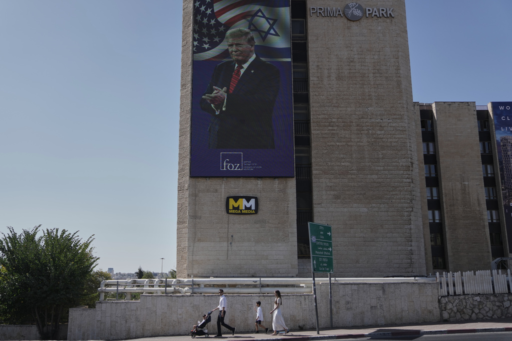 People walk past large outdoor screen displaying an image depicting the U.S. President Donald Trump, in Jerusalem, Saturday, Oct. 11, 2025, after Israel and Hamas agreed to a pause in their war and the release of the remaining hostages. (AP Photo/Mahmoud Illean)
