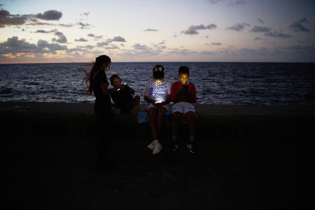 Children look at their phones while sitting on the Malecón wall during a blackout in Havana, Cuba, Saturday, March 21, 2026. (AP Photo/Ramon Espinosa)