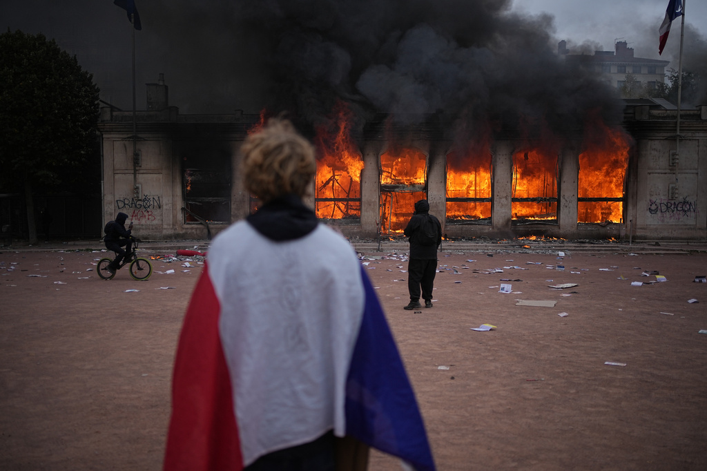 A protester wraps a French flag around them in front of the burning tourism office during a demonstration against rising living costs and in solidarity with students&rsquo; grievances in Lyon, France, Saturday, Oct. 17, 2026. (AP Photo/Laurent Cipriani)