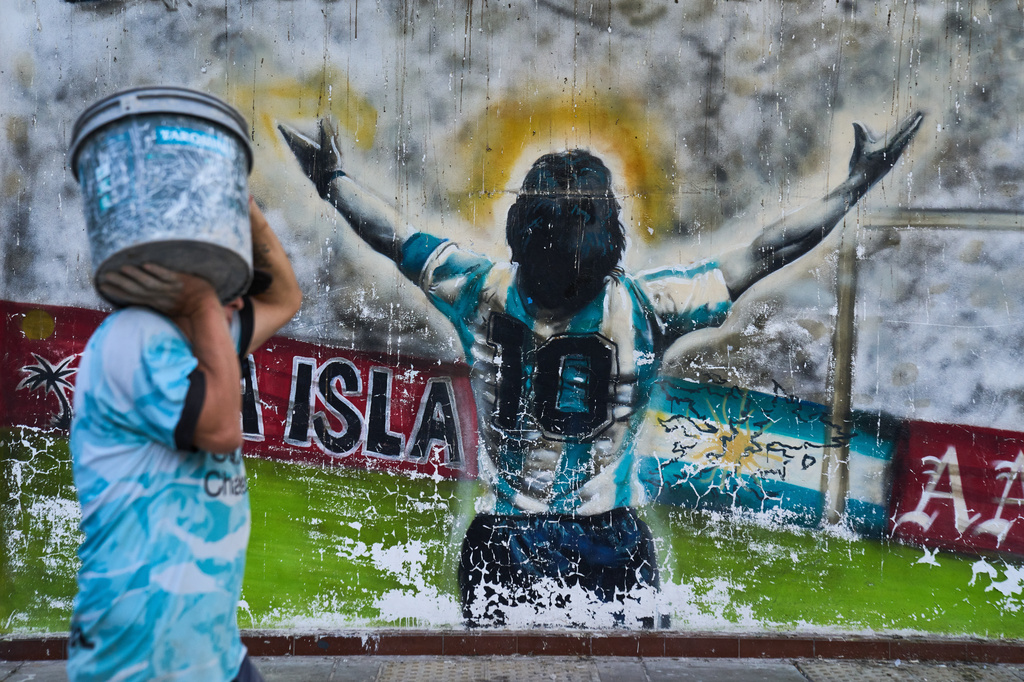 A pedestrian carries a bucket past graffiti depicting late soccer legend Diego Maradona in Buenos Aires, Argentina, Monday, April 13, 2026. (AP Photo/Rodrigo Abd)