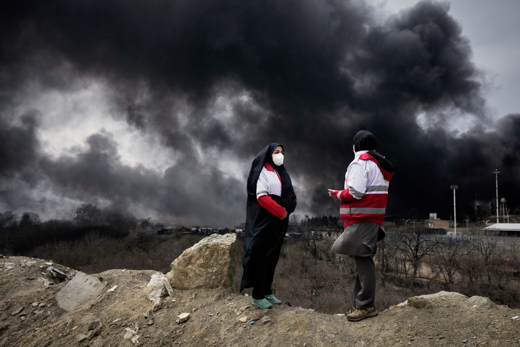 Two women from the Iranian Red Crescent Society stand as a thick plume of smoke from a U.S.-Israeli strike on an oil storage facility late Saturday rises in the sky in Tehran, Iran, Sunday, March 8, 2026. (AP Photo/Vahid Salemi)