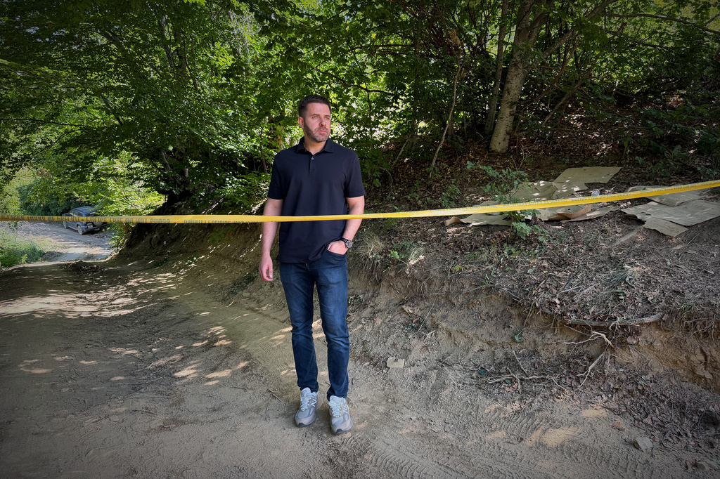 Erblin Ademi, whose father, Adem Ademi, a prominent surgeon, disappeared along with 22 other intellectuals during the Kosovo war in April 1999, stands behind yellow tape near the excavation site of a mass grave in Small Kalluder, Kosovo, Wednesday, Aug. 5, 2026. (AP Photo/Zana Cimili)