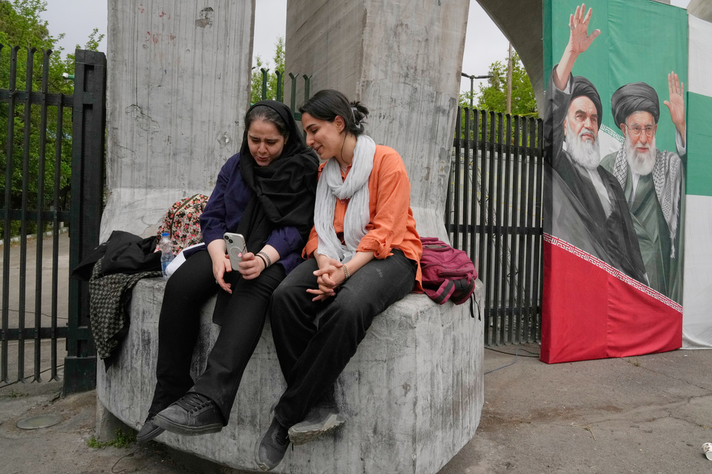 Women share a moment as they look at a smartphone at the main gate of the Tehran University as a banner shows portraits of the late Supreme Leader Ayatollah Ali Khamenei, right, and the late revolutionary founder Ayatollah Khomeini in Tehran, Iran, Sunday, April 19, 2026. (AP Photo/Vahid Salemi)
