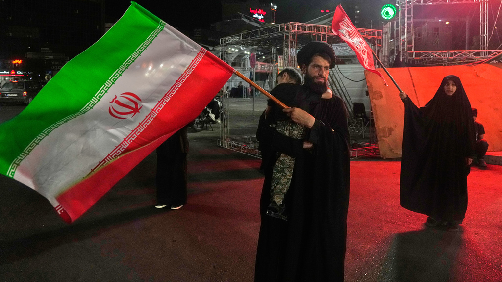 A cleric waves an Iranian flag while holding a child as a woman waves a religious flag in a pro-government gathering commemorating the late Iranian Supreme Leader Ayatollah Ali Khamenei at a square in Tehran, Saturday, July 11, 2026. (AP Photo/Vahid Salemi)