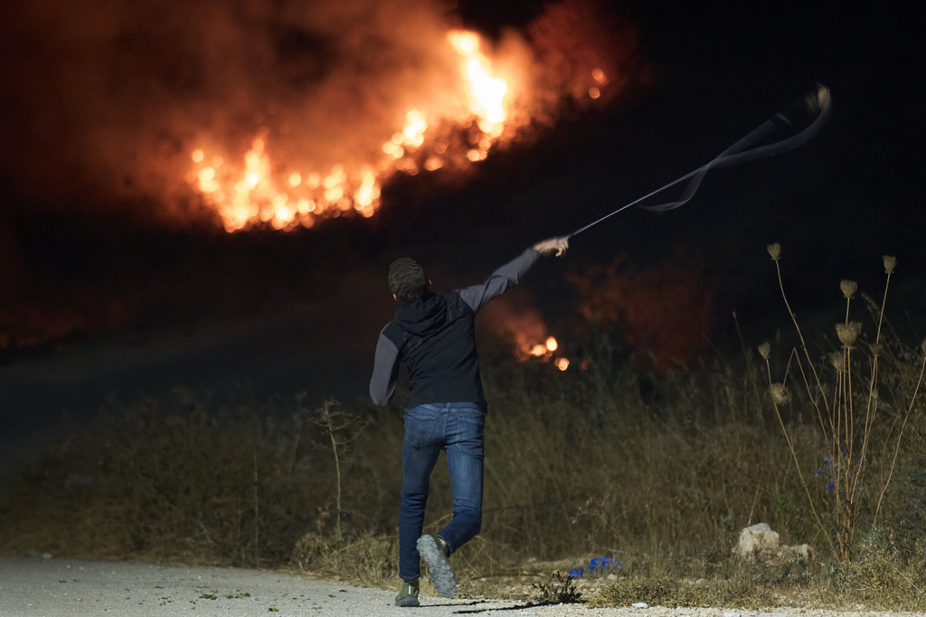 A Palestinian uses a slingshot during clashes with Israeli settlers as brush burns after it was set ablaze to prevent them from approaching the West Bank village of Sinjil, Friday, July 17, 2026. (AP Photo/Leo Correa)