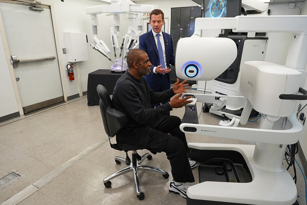 Colorado head football coach Deion Sanders, front, reacts after working the Da Vinci robotic surgical machine used to remove his cancerous bladder last year during a demonstration of the device by Jason Hart, senior vice president of global marketing for Intuitive, Tuesday, June 23, 2026, in Boulder, Colo. (AP Photo/David Zalubowski)