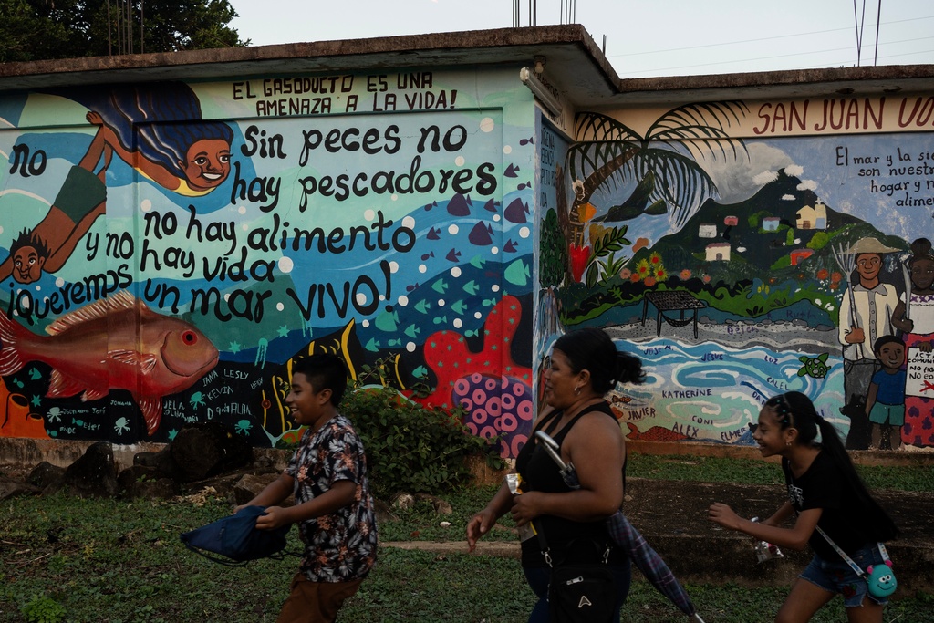 A family walks past a mural defending the territory with messages like "The gas pipeline is a threat to life!" and "We want a living sea!" in San Juan Volador, Mexico, Oct. 27, 2025. (AP Photo/Felix Marquez)