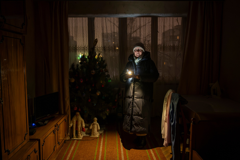 Lyudmila Kopal wearing warm clothes shines a flashlight as she stands by a Christmas tree during a blackout caused by Russia's regular air attacks against the country's energy in Kyiv, Ukraine, Tuesday, Jan. 13, 2026. (AP Photo/Danyil Bashakov)