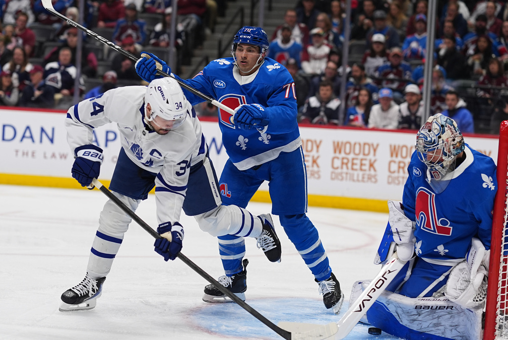 Toronto Maple Leafs center Auston Matthews, left, directs the puck at Colorado Avalanche goaltender Trent Miner, right, while being checked by Avalanche defenseman Sam Malinski, second from left, in the second period of an NHL hockey game Monday, Jan. 12, 2026, in Denver. (AP Photo/David Zalubowski)