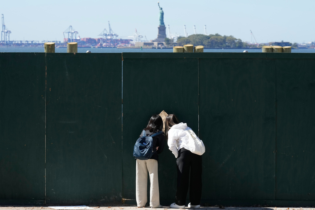 FILE - People look through an opening in a fence to get a glance at the Statue of Liberty in New York, Oct. 1, 2025. (AP Photo/Seth Wenig, File)