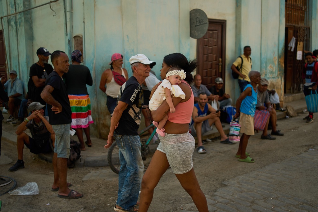 A woman walks with a baby in her arms past people lined up to buy bread during a blackout in Havana, Cuba, Thursday, March 5, 2026. (AP Photo/Ramon Espinosa)
