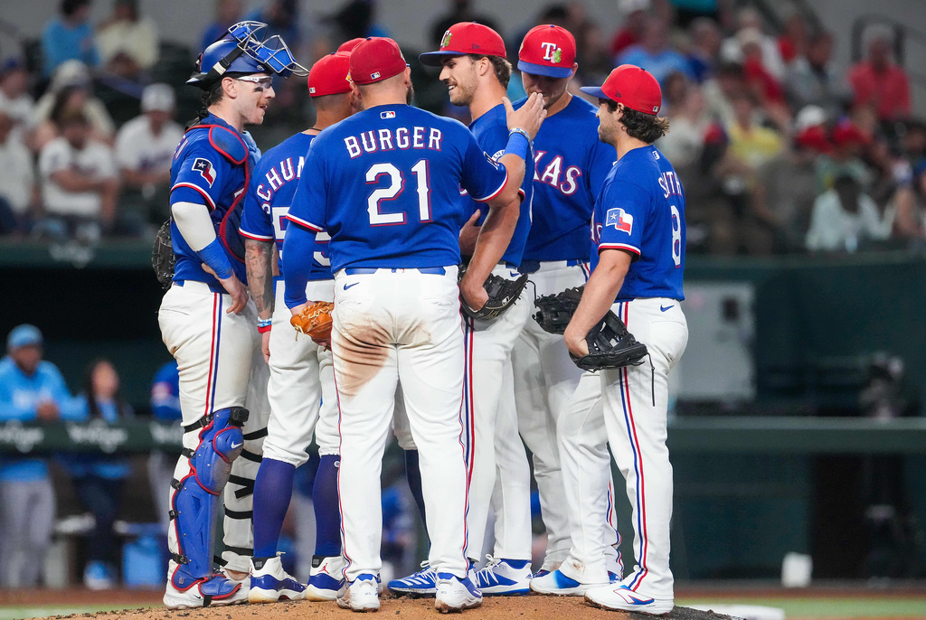 Texas Rangers pitcher Carter Baumler, third from right, gets a pat on the back from first baseman Jake Burger (21) as he gets a visit at the mound from manager Skip Schumaker, second from left, to inform him he has made the opening day roster during the fifth inning of an exhibition baseball game against the Kansas City Royals, Monday, March 23, 2026, in Arlington, Texas. (Smiley N. Pool/The Dallas Morning News via AP)