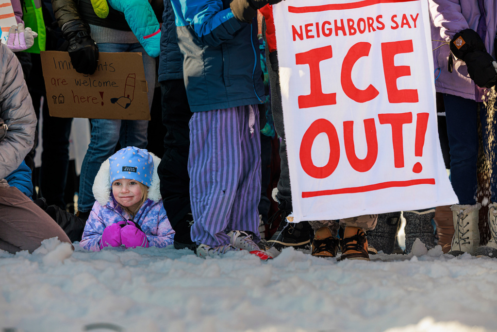 A child sits in the snow as demonstrators hold signs during a news conference at Lake Hiawatha Park in Minneapolis, on Friday, Jan. 9, 2026, demanding Immigration and Customs Enforcement be kept out of schools and Minnesota following the killing of 37-year-old mother Renee Good by federal agents earlier on Wednesday. (Kerem Yücel/Minnesota Public Radio via AP)