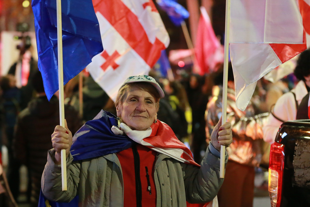 A demonstrator holds a Georgian and an EU flag as others gather in the Georgian capital of Tbilisi, on Friday, Nov. 28, 2025, to mark 365 days of non-stop protests against the government's decision to halt talks on joining the European Union. (AP Photo/Zurab Tsertsvadze)